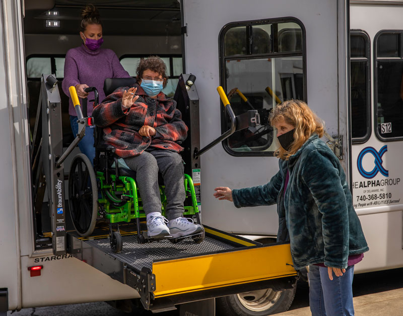 The Alpha Group Transportation, masked person in wheelchair smiling and waving while being helped off transportation